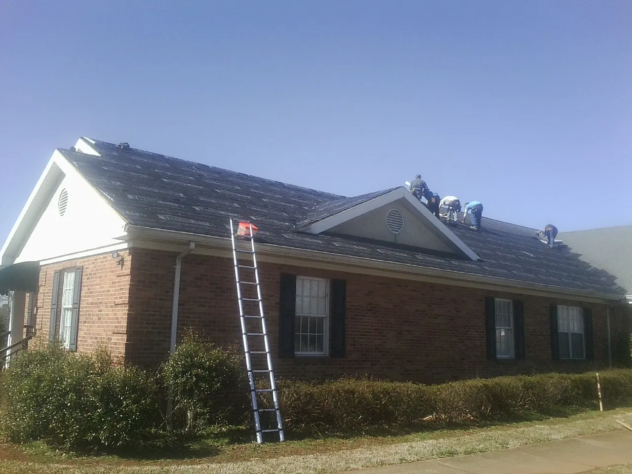 Crew tearing shingles off a brick ranch roof with a ladder set — Emergency Roof Repair work by our crew in Hockessin