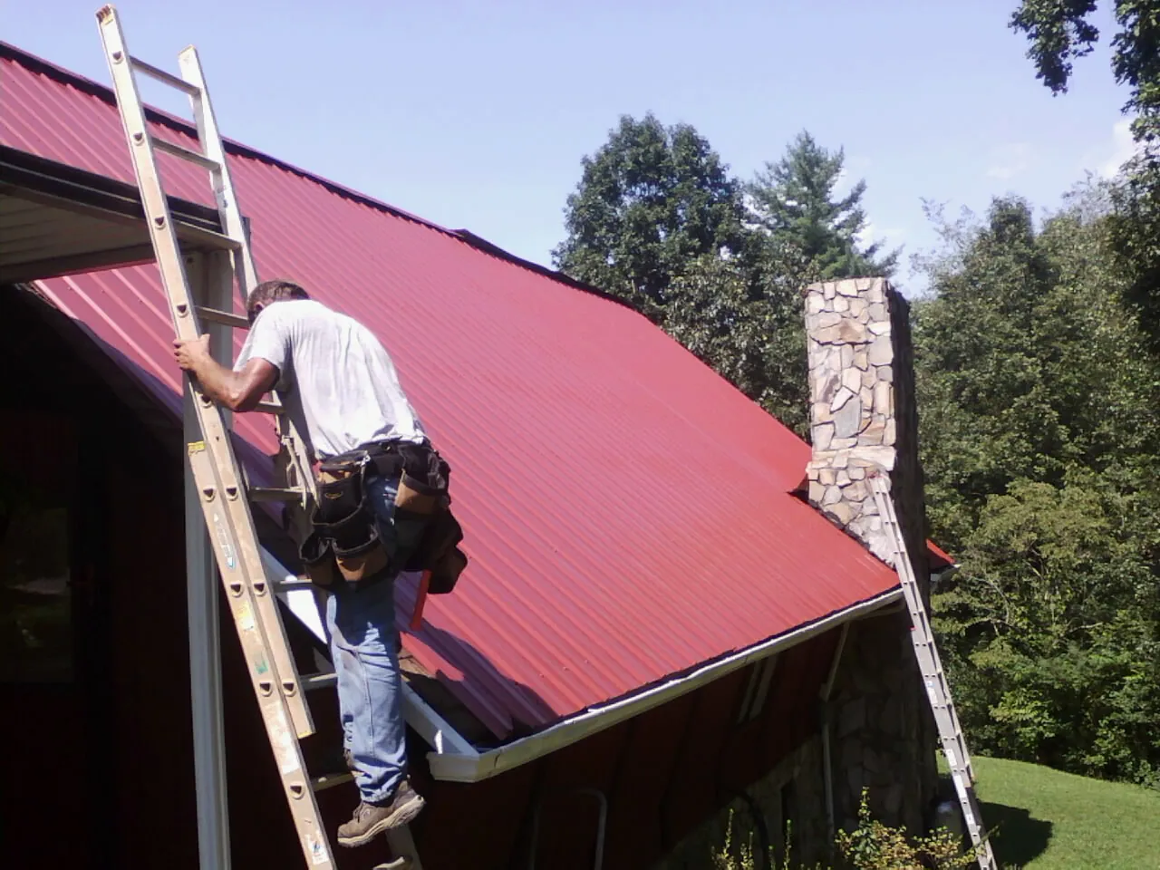 Roofer on a ladder beside a red metal roof and stone chimney — Soffit & Fascia Repair by Roofing Brothers in Hockessin